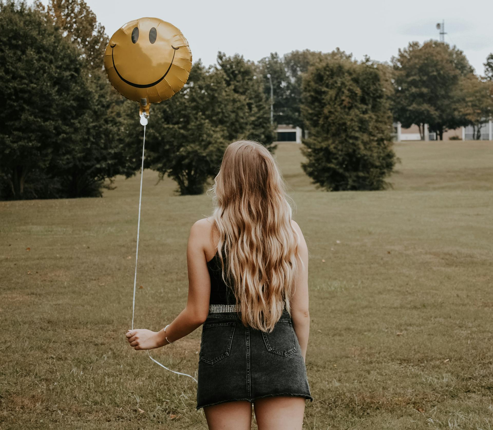 A woman with long hair holds a smiley balloon in a green park, embracing nature and positivity.