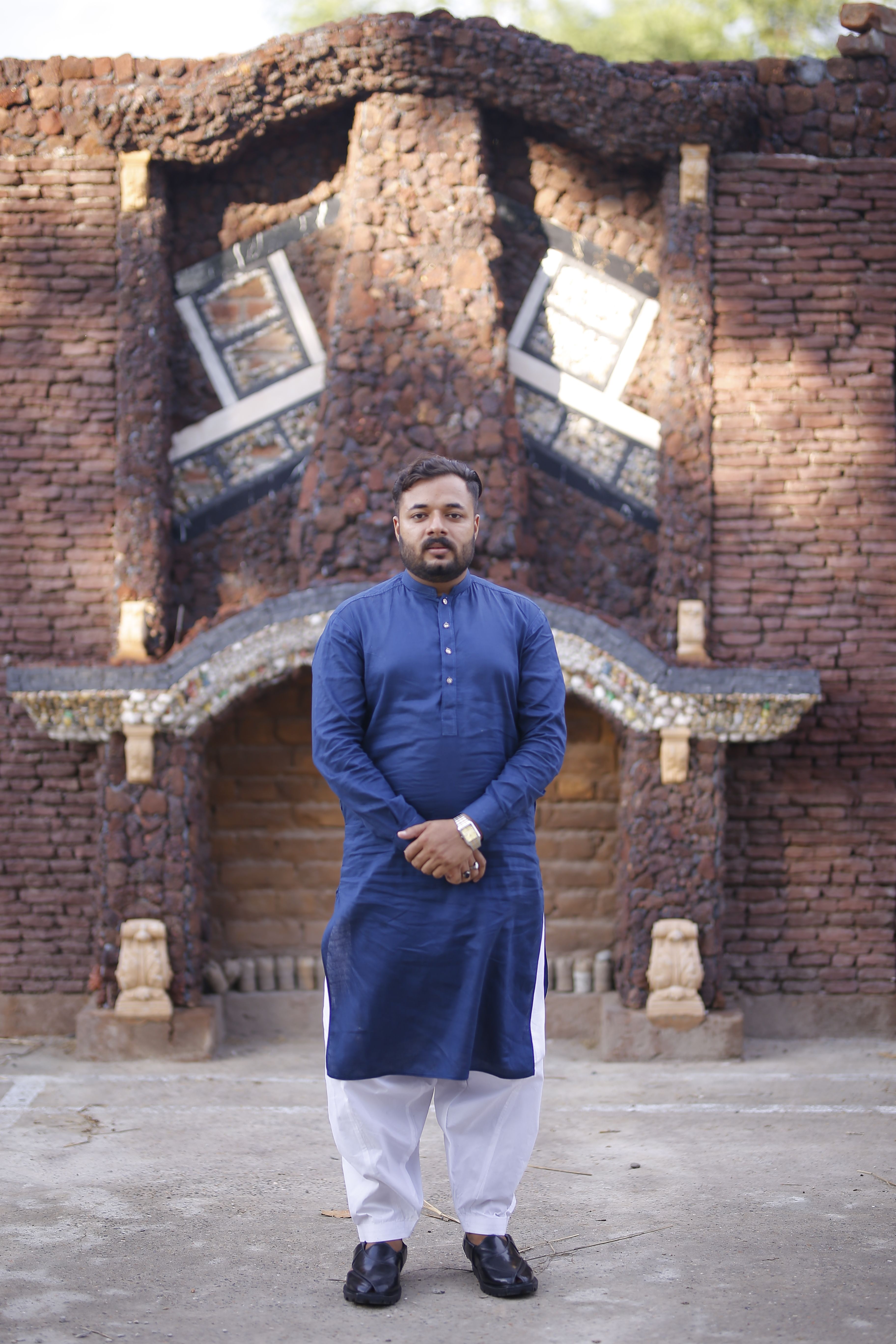 Asian man in blue traditional clothes standing in front of a rustic brick wall in Pakistan.