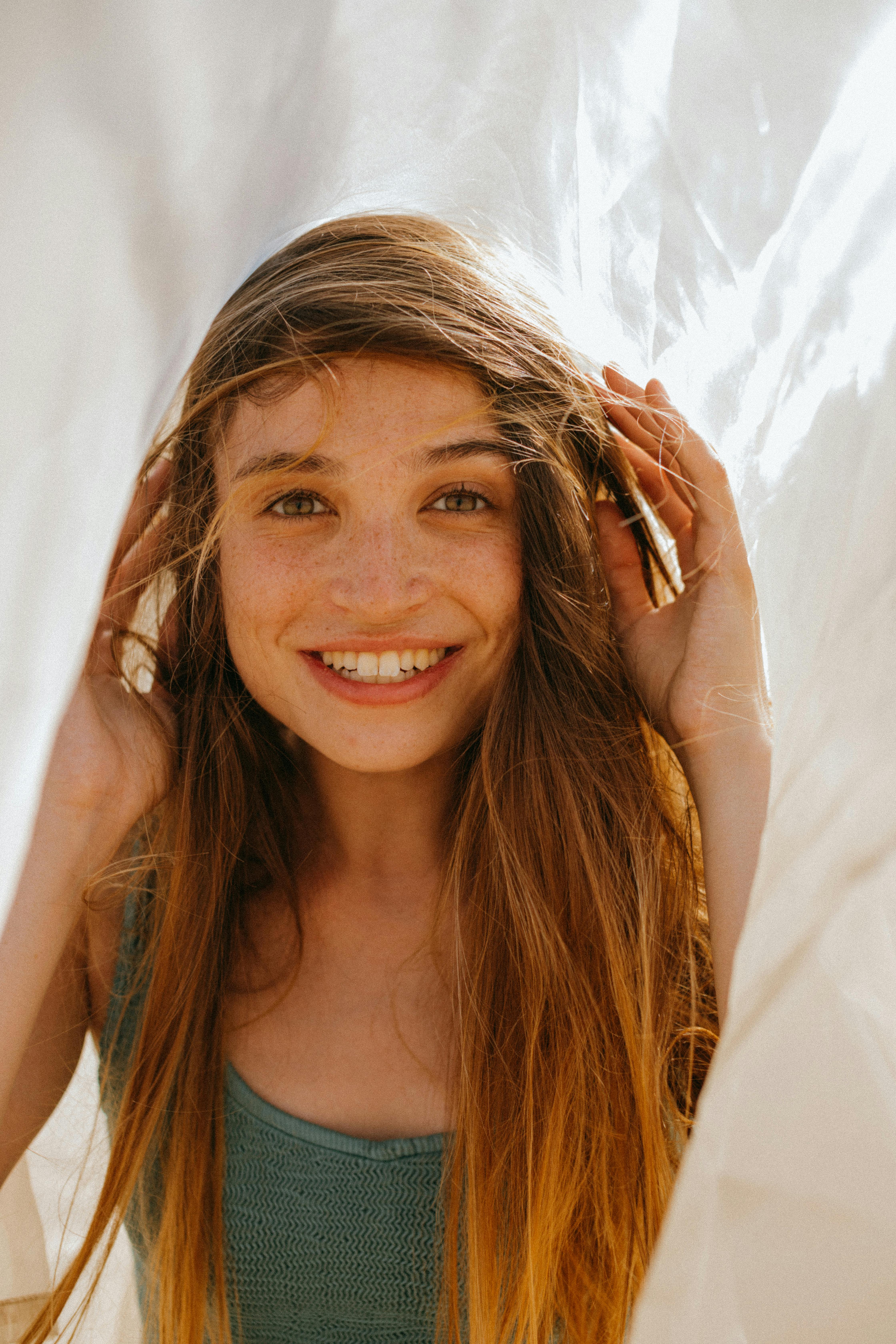 A cheerful woman with long hair smiling warmly outdoors under sunlight.
