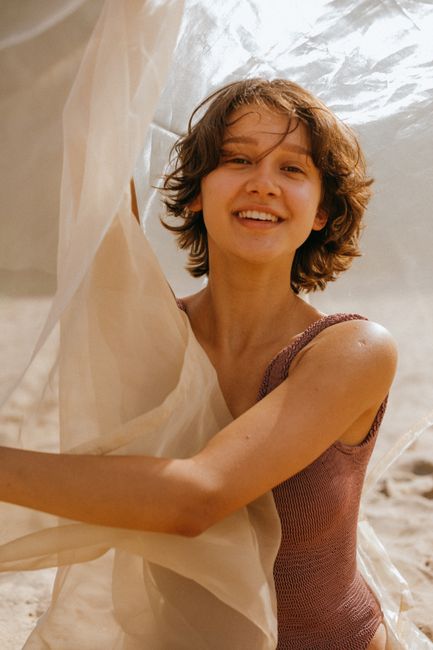A smiling woman in a swimsuit plays with sheer fabric on a sunny beach day.