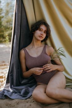 Young woman in a one-piece swimsuit sits holding leaves, with draped fabric backdrop.