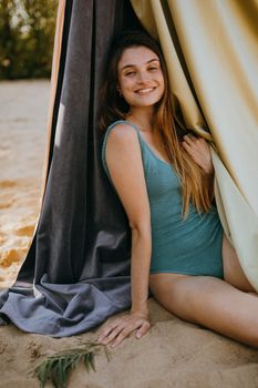 Caucasian woman in a teal swimsuit sitting on a sandy beach, smiling warmly.