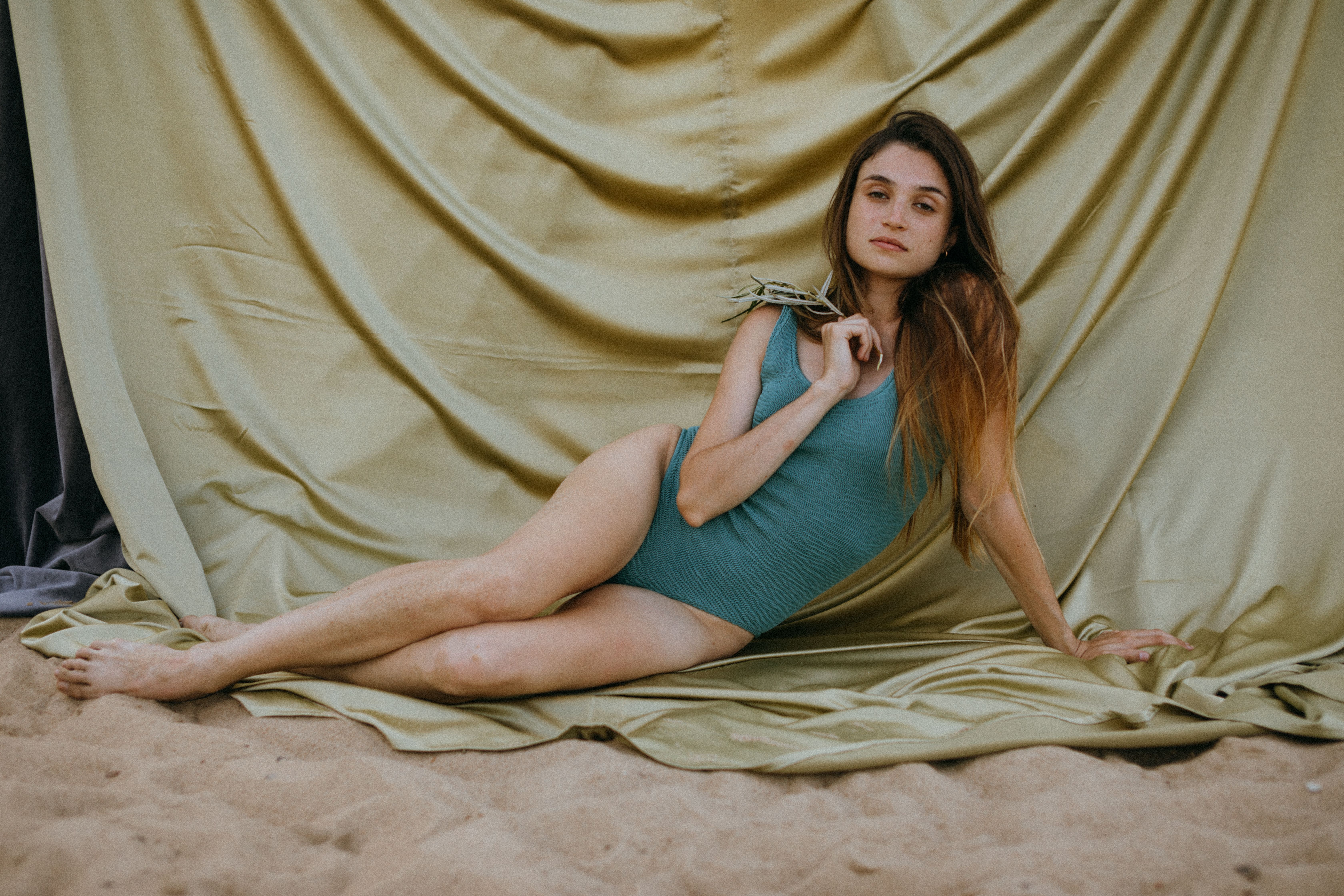 Model in a teal swimsuit lounging on sand with draped silk backdrop.