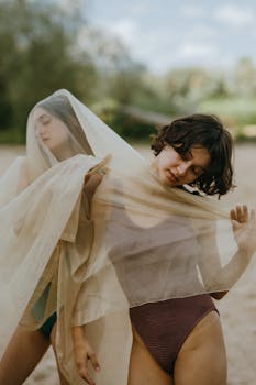 Two women in bodysuits dance gracefully with sheer fabric in a natural outdoor setting.