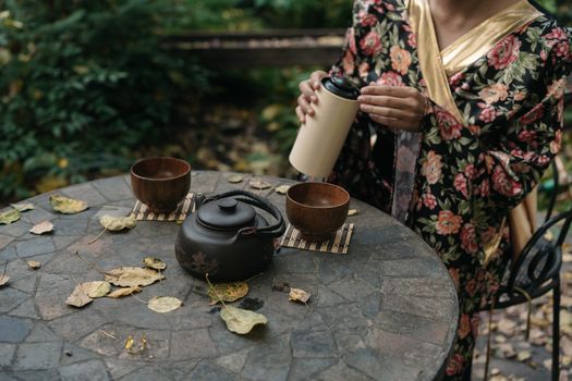 A woman in a floral kimono prepares tea in a serene garden setting.