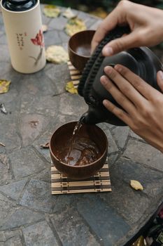 Hands pouring tea from a teapot into a wooden bowl on a stone table outdoors.