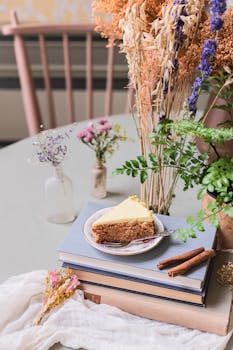 Slice of cake with cinnamon sticks on books, surrounded by wildflowers indoors.