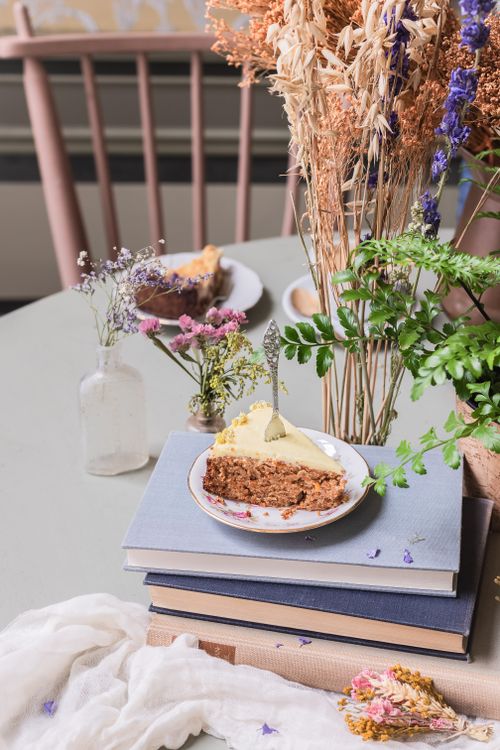 Free A slice of carrot cake with frosting sits on stacked books, surrounded by dried flowers and rustic decor. Stock Photo