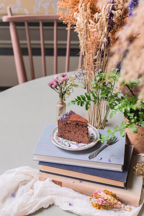 Free A slice of chocolate cake on a stack of books, surrounded by dried flowers in a cozy cafe setting. Stock Photo