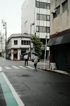 Urban street scene on a rainy day with people walking under umbrellas, among tall buildings.
