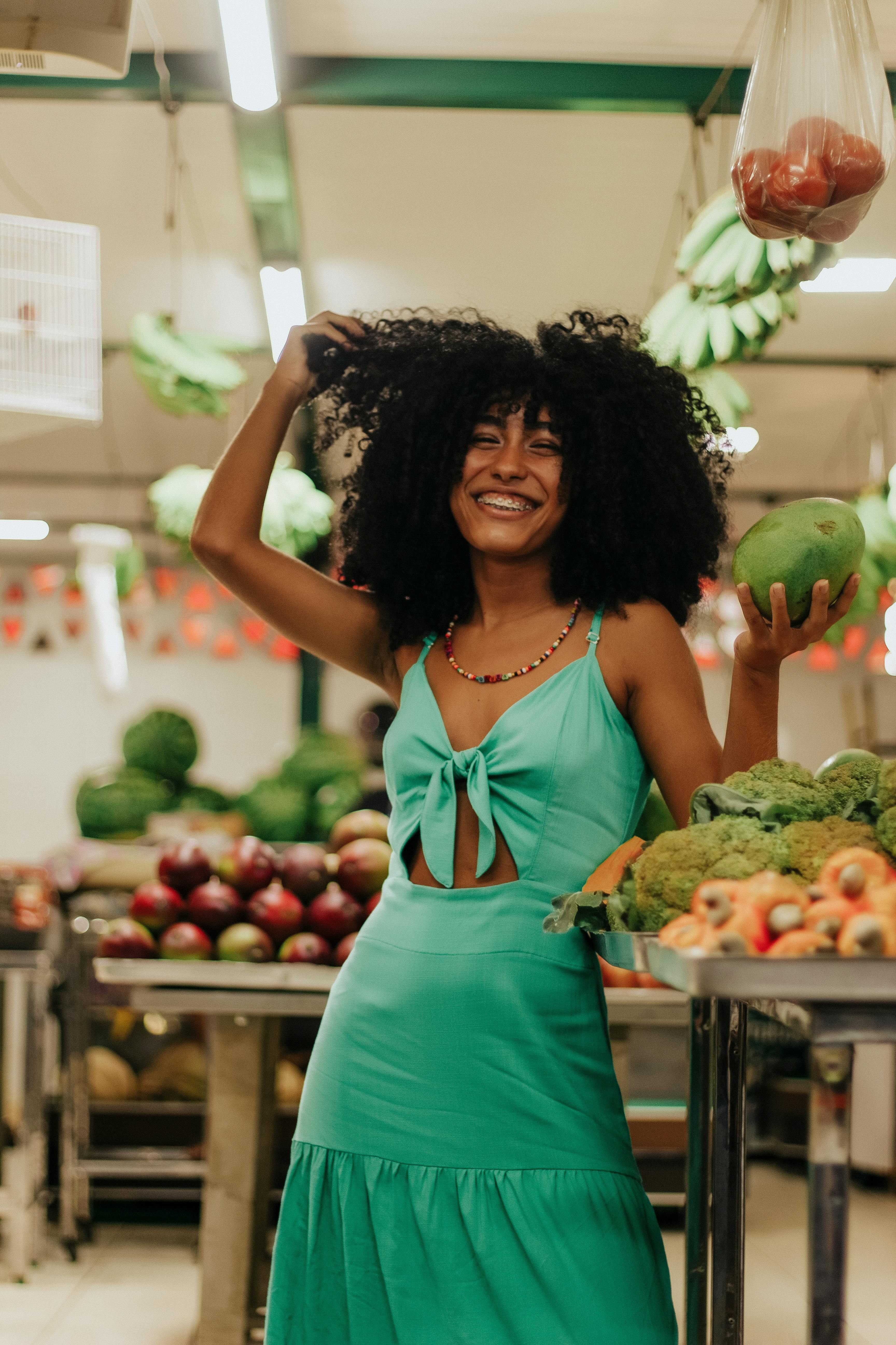 Joyful woman with afro hair posing with fruit at a vibrant Brazilian market.