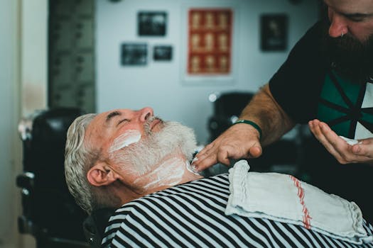 Close-up of a barber applying shaving cream to an adult male's beard in a barbershop.