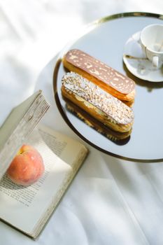 Two gourmet eclairs on a mirrored tray with a book and a peach, creating a serene and indulgent setting.
