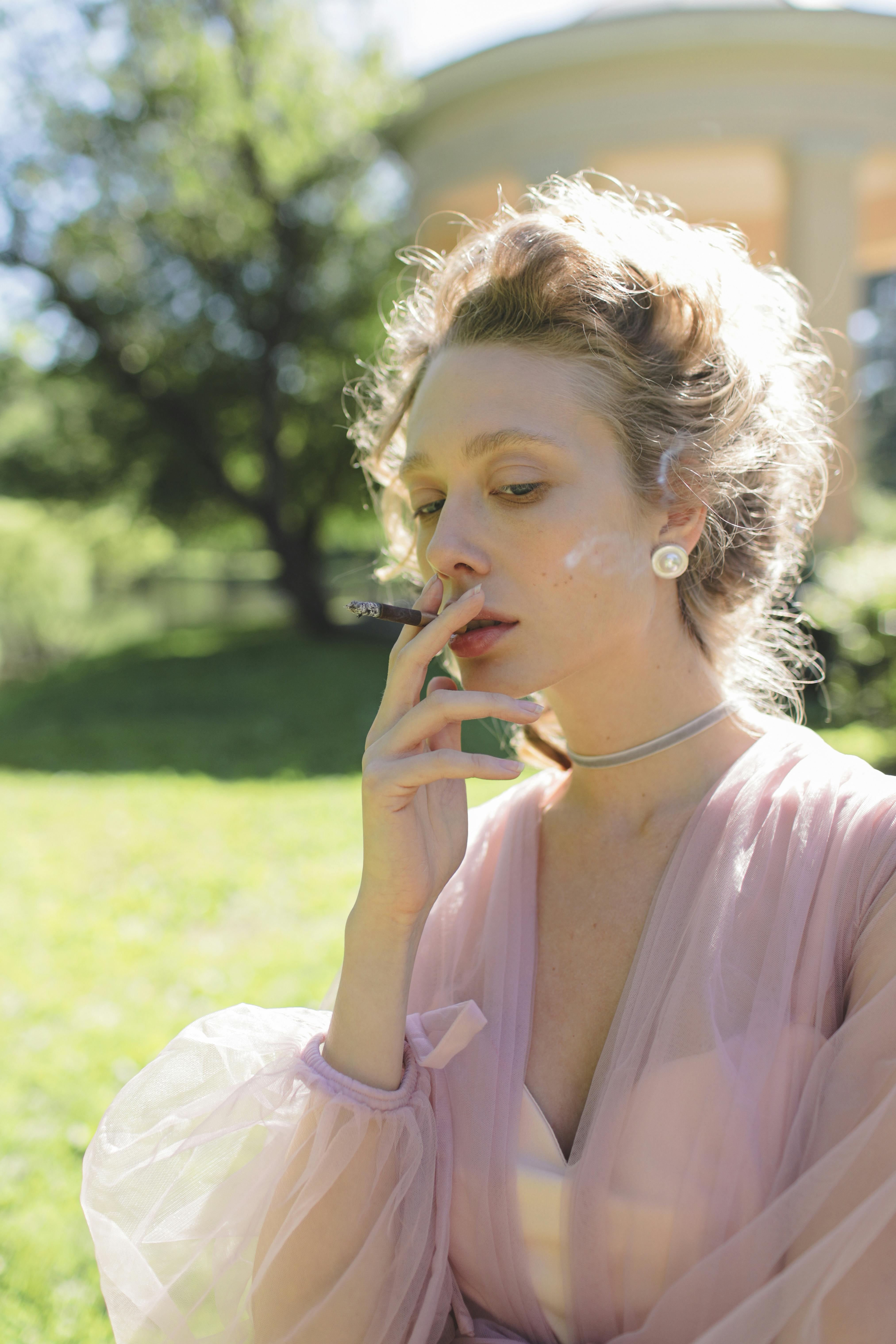 Graceful woman in a pink dress smoking in a sunlit garden setting.