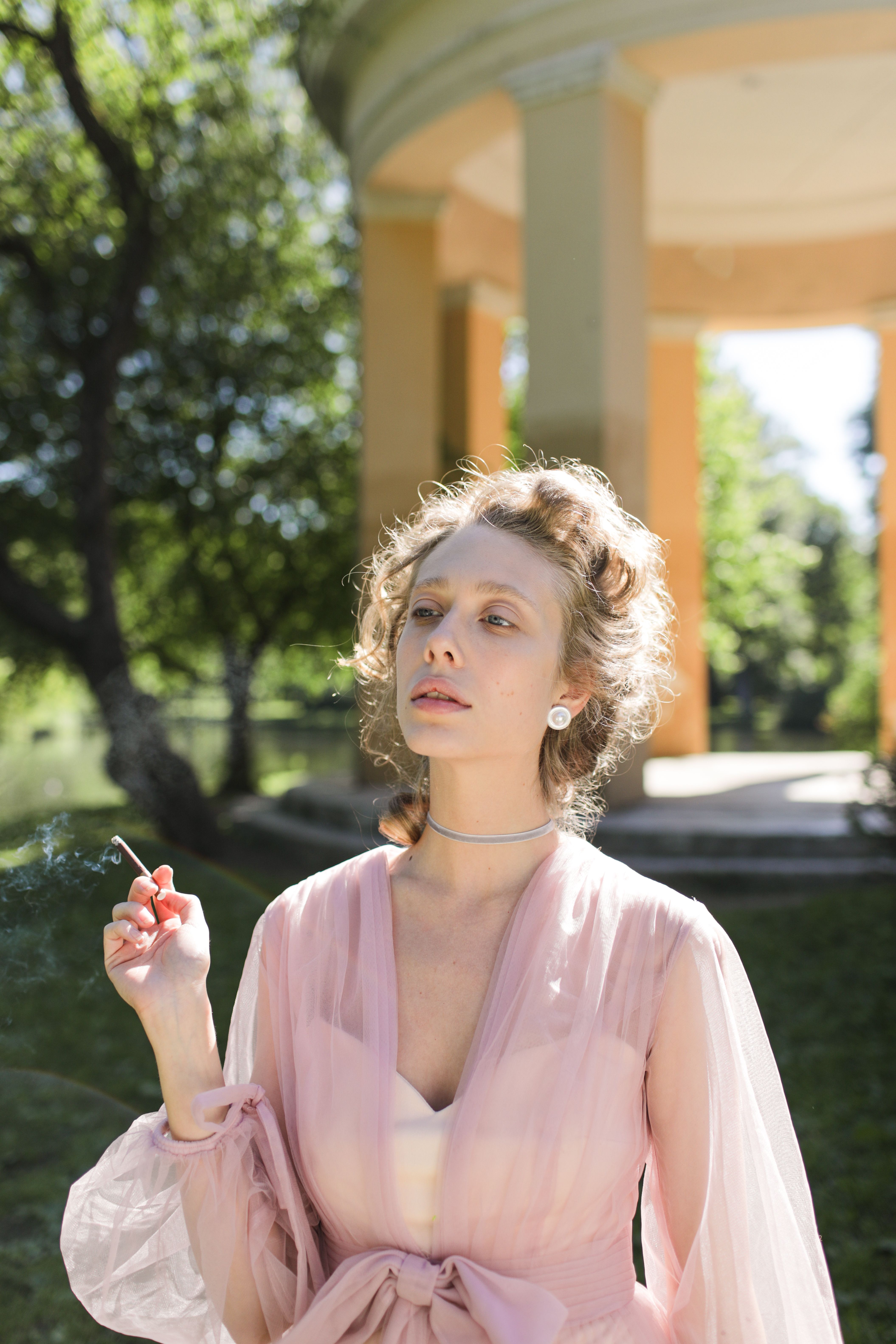 Elegant woman in a pink dress, smoking in a serene outdoor setting.