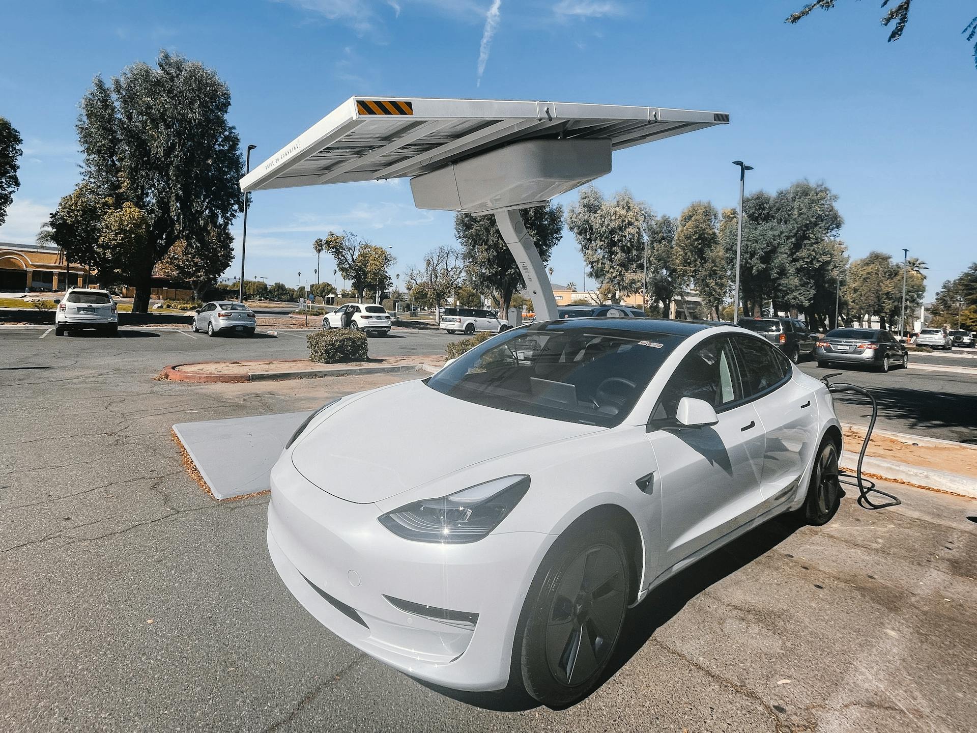 Modern white electric car charging under solar panels in a sunny parking lot.