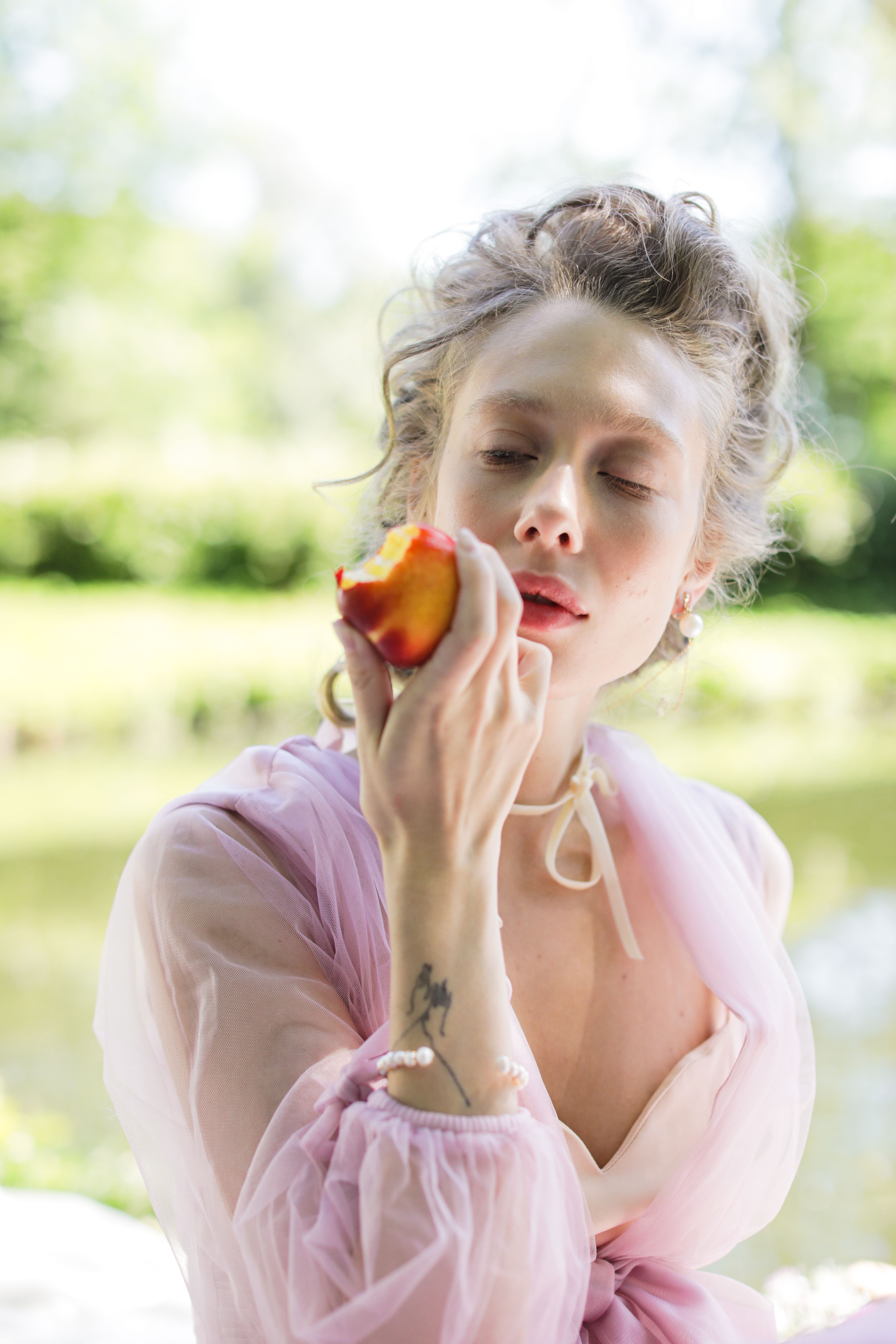 Woman with tattoo enjoying a peach in a serene outdoor setting.