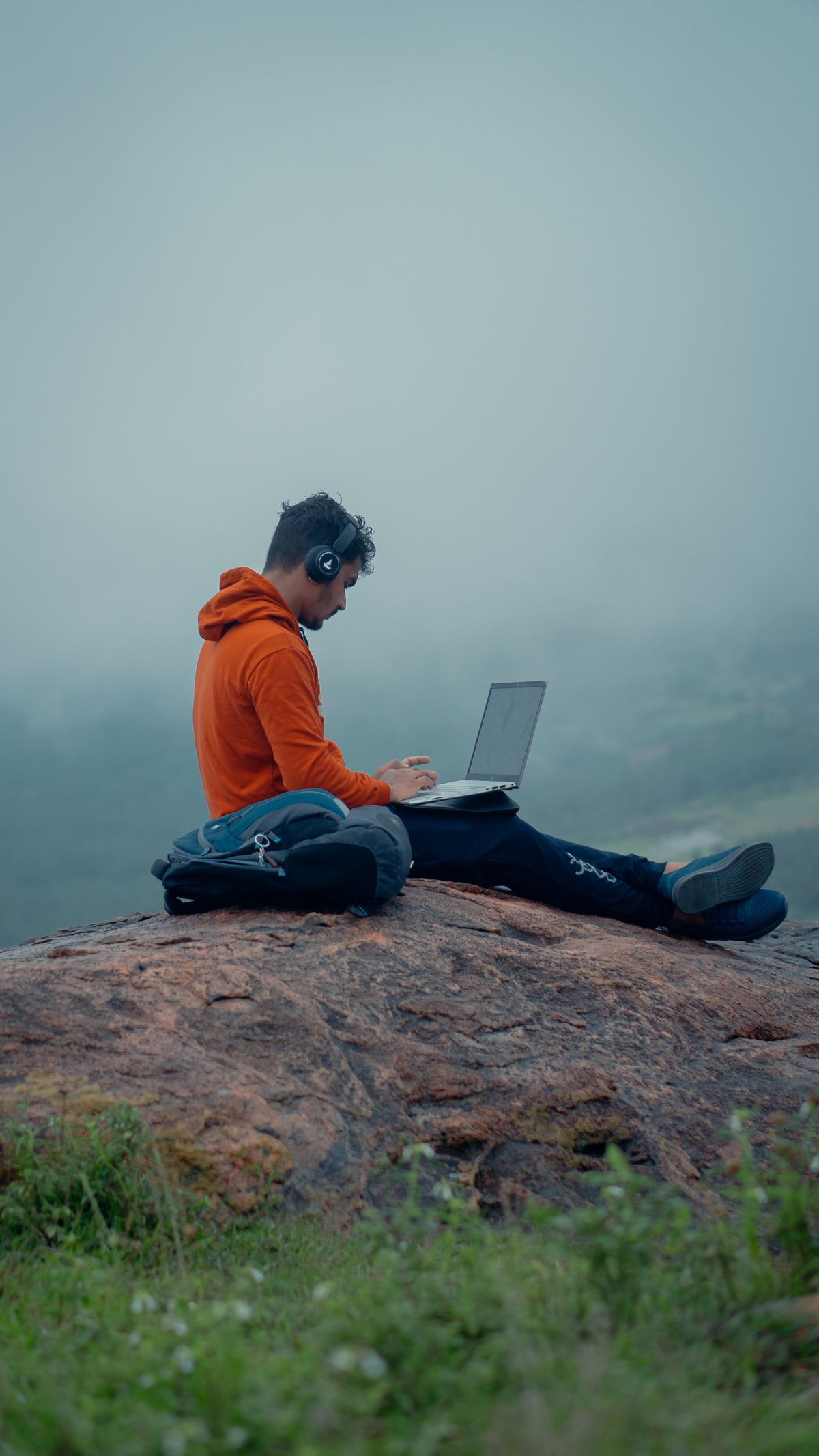 Pria pakai laptop dan headphone bekerja remote di alam terbuka