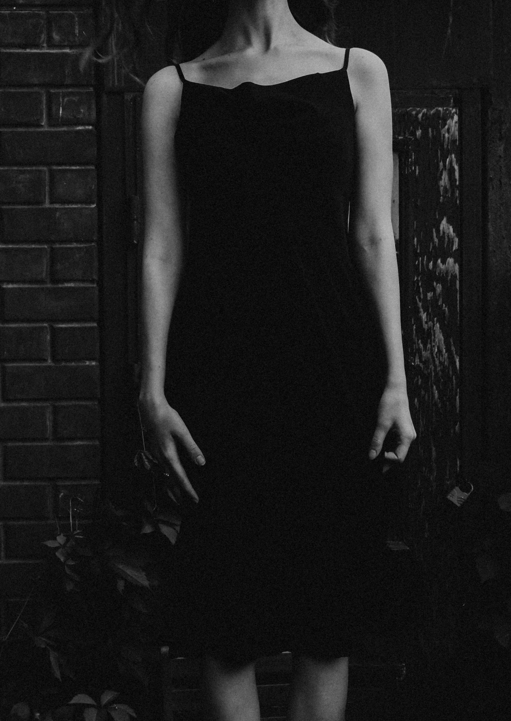 Black and white photo of a woman in an elegant black dress standing against a textured brick wall.