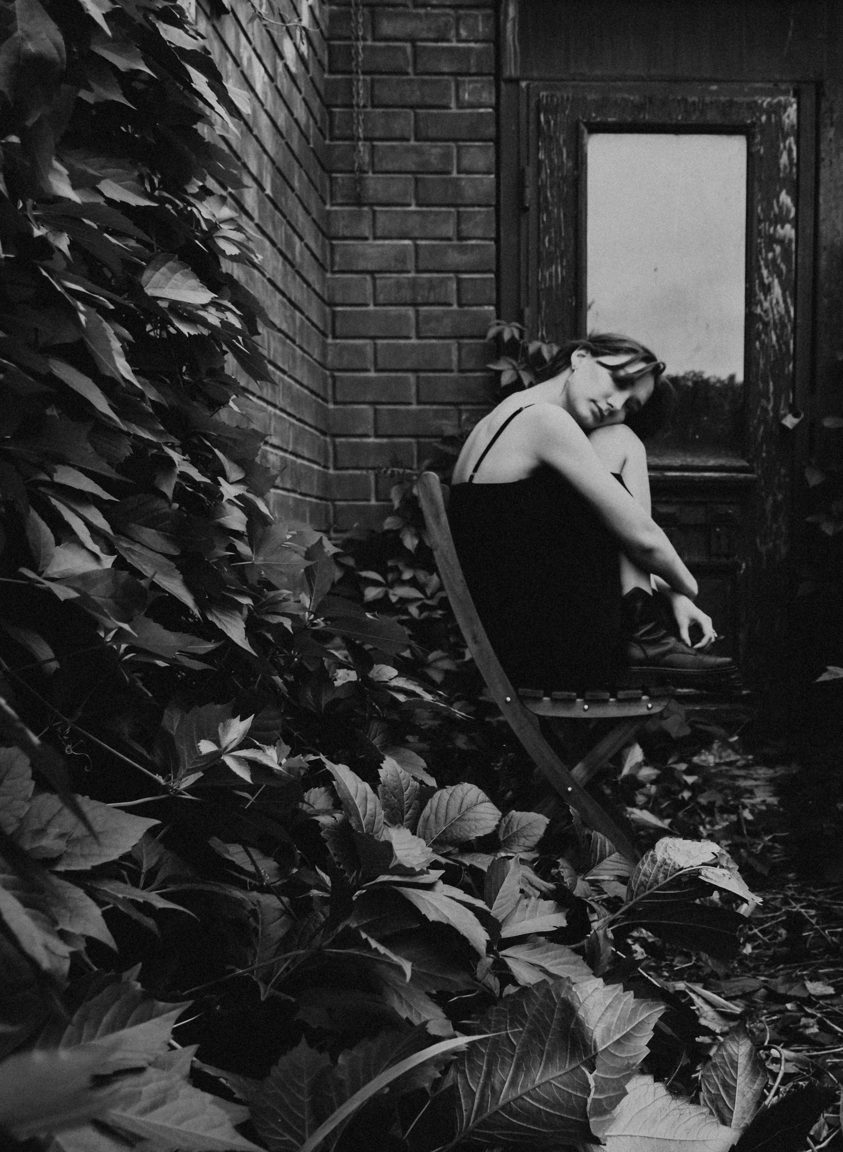 Black and white image of a woman sitting on a chair outdoors near a brick wall and door.