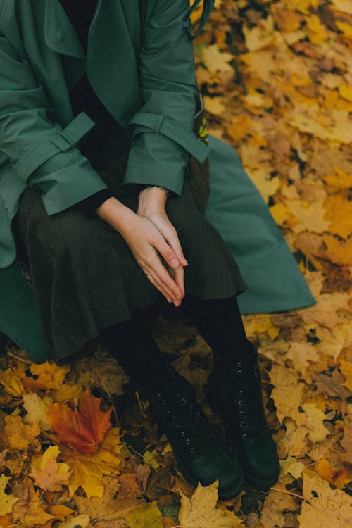 Young woman in green trench coat sitting on ground covered with autumn leaves