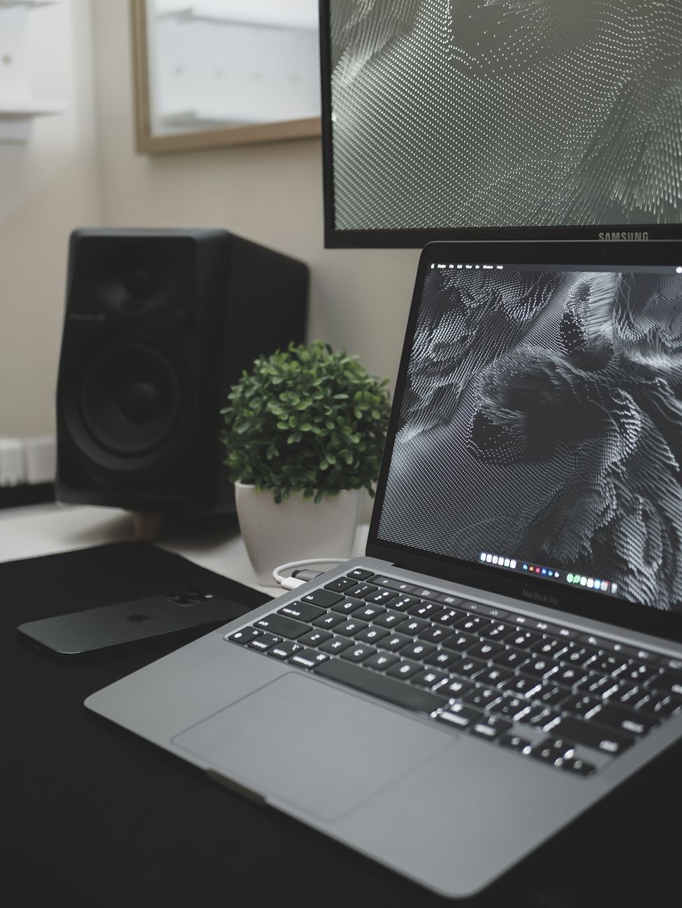 A sleek workspace featuring a laptop with digital art, monitor, speaker, and potted plant.