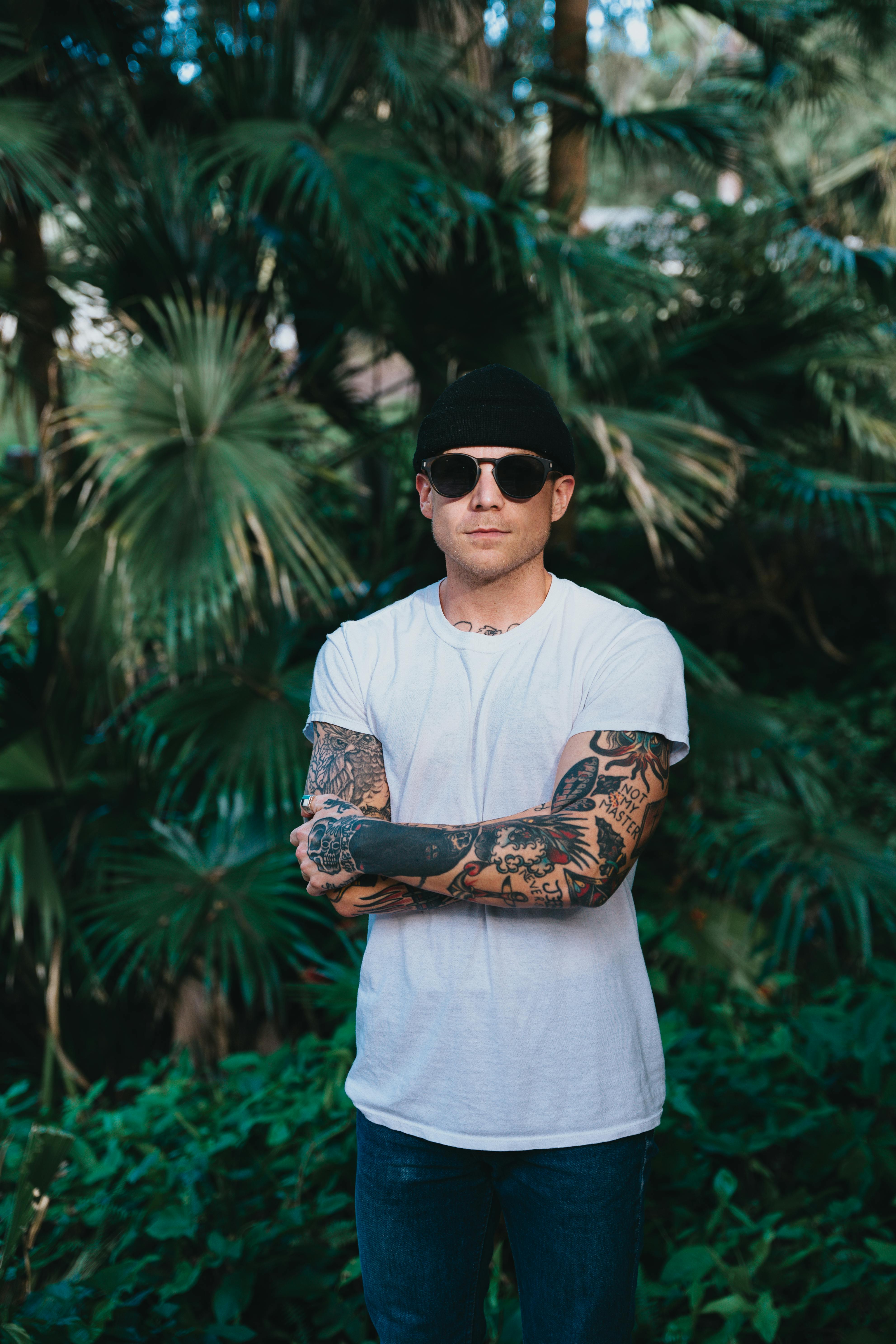 Man in sunglasses and tattoos poses confidently in front of lush greenery.