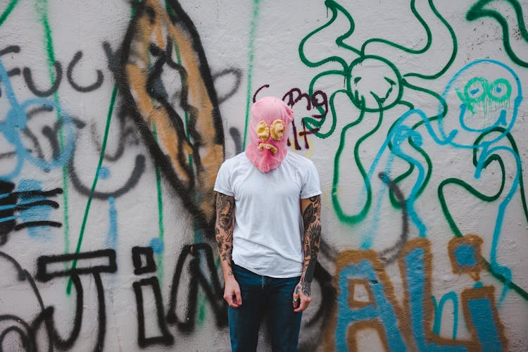 Man In Pink Bag Over His Head With Sad Face Sewn On Standing In Front Of Graffiti