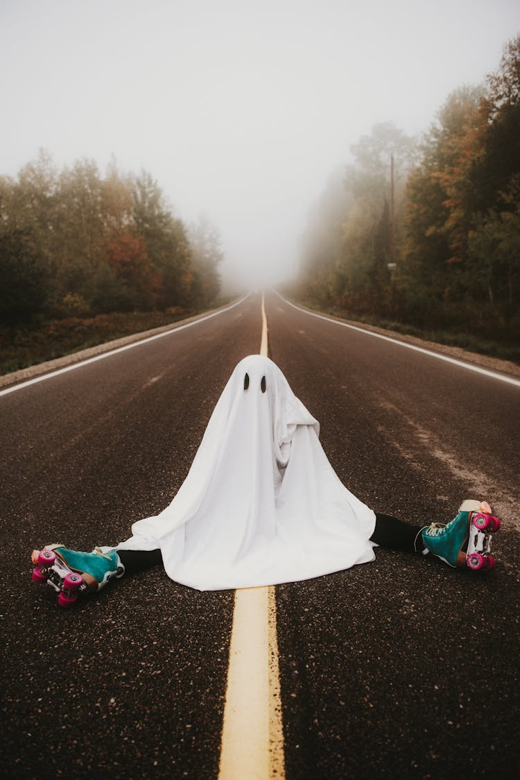 Person In Ghost Costume And Roller Skates Sitting On Foggy Road