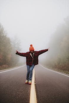 Woman stands with arms outstretched on a foggy road, creating symmetry amidst misty trees.