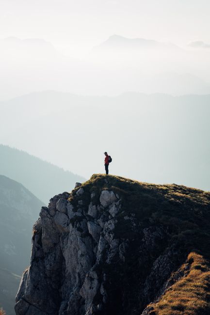 Lone hiker in red jacket standing on a steep mountain cliff with misty mountains in background.