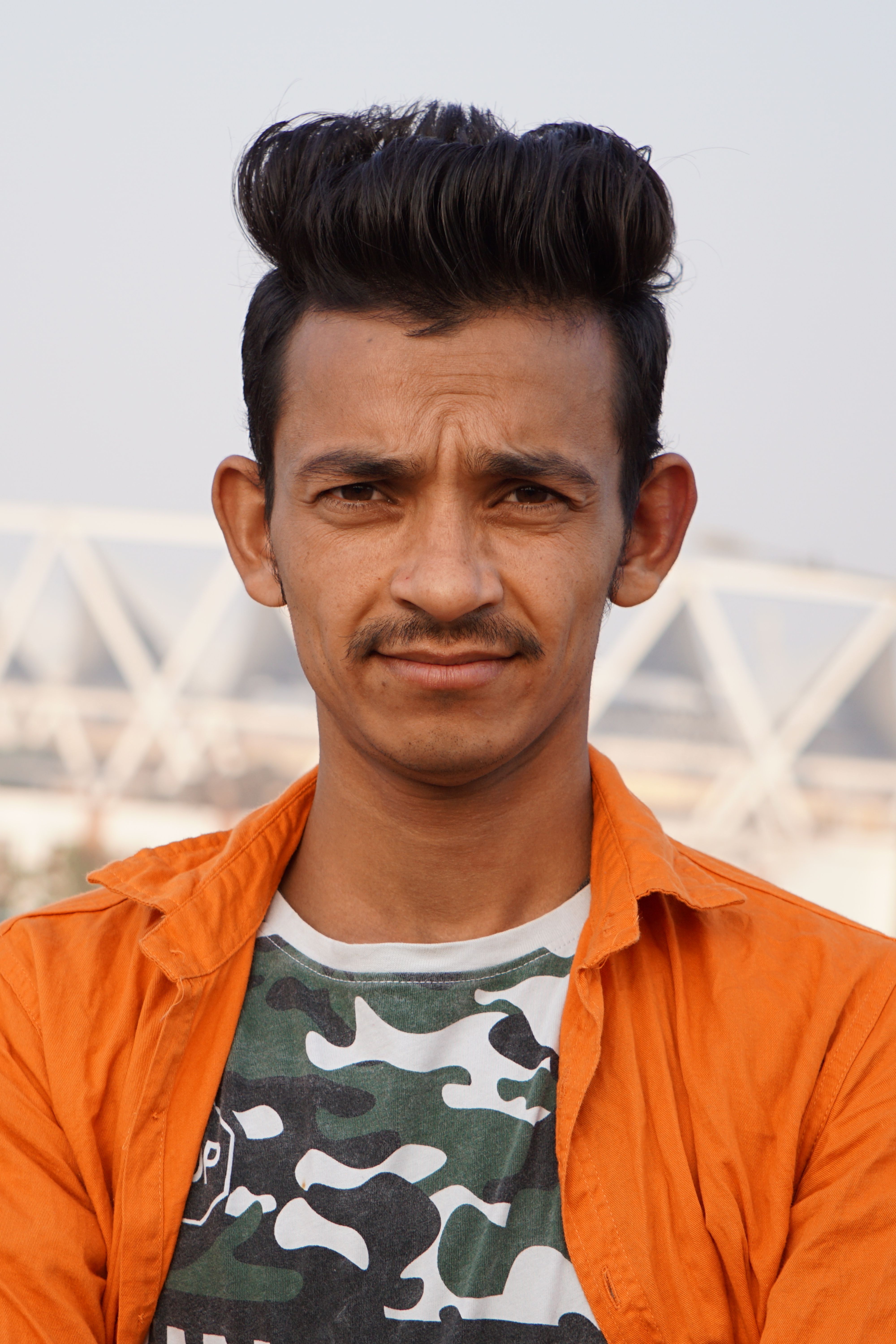 Close-up portrait of a man with distinctive hairstyle and mustache in an outdoor setting.