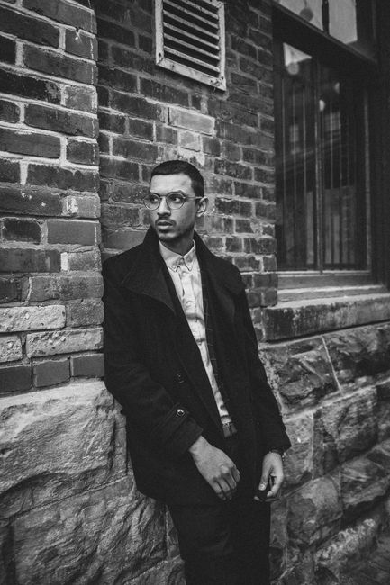 Black and white portrait of a man posing against an urban brick wall outdoors.