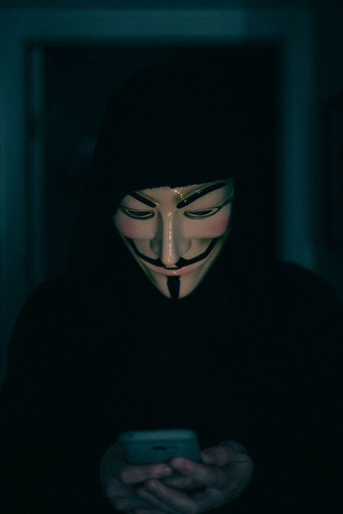 Osoba s Guy Fawkes maskom i tamnom kapuljacom gleda u smartphone, simbolizirajući anonimnost i privatnost na internetu