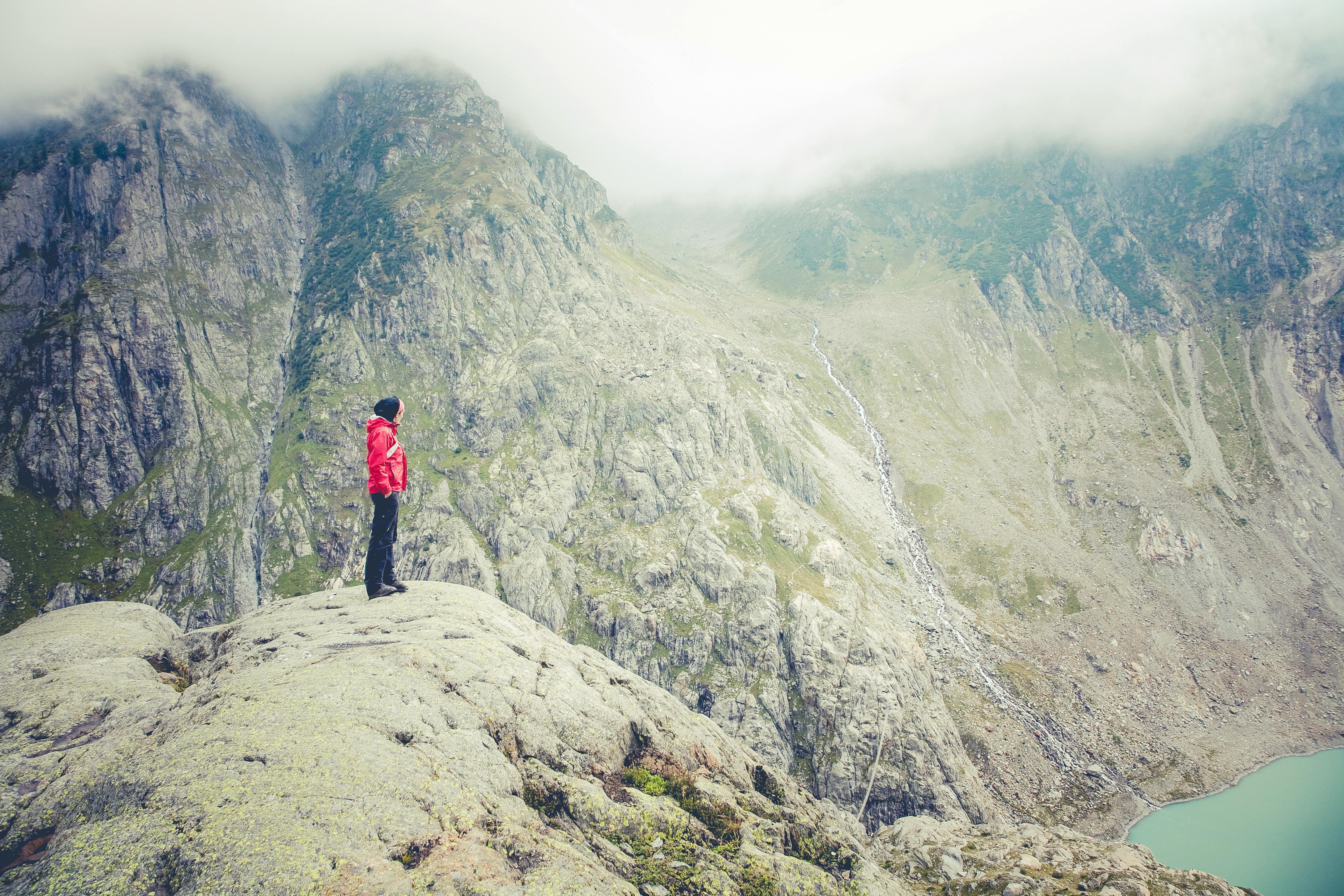 一名穿着红色夹克的人在大雾天气中，从岩石山上俯瞰宁静的高山湖泊，欣赏着令人惊叹的景色