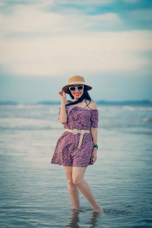 Free A cheerful woman in a floral dress enjoying a sunny beach day, wearing a hat and sunglasses. Stock Photo