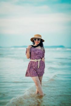 Asian woman in a floral dress with a hat posing by the sea on a sunny day.