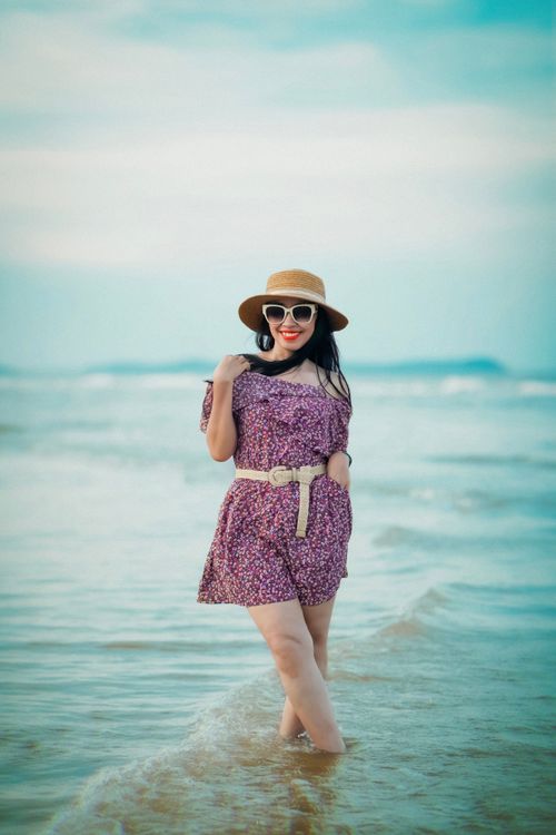 Free Asian woman in a floral dress with a hat posing by the sea on a sunny day. Stock Photo