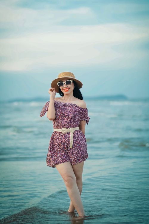 Free Woman in a floral dress, hat, and sunglasses enjoying a beach day by the sea. Stock Photo