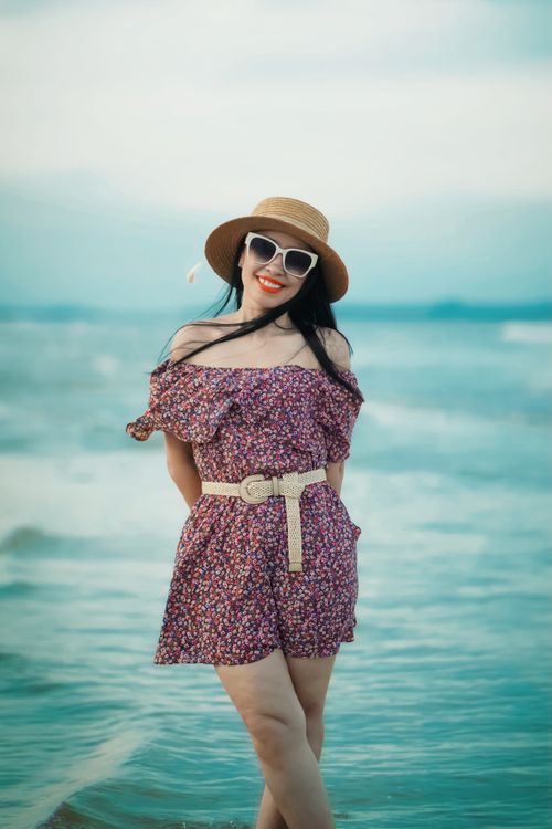 Free Elegant woman in floral dress and hat posing with a smile on a sunny beach day. Stock Photo