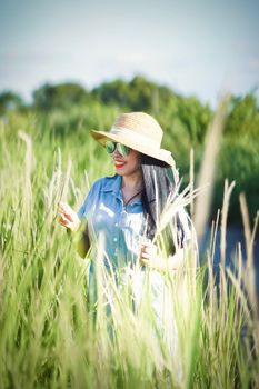 A woman with a straw hat and sunglasses smiles among tall grass during a sunny summer day.