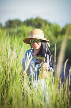 A fashionable woman with a straw hat and sunglasses smiling in a green field under bright sunlight.