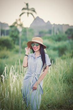 A fashionable woman in a hat and sunglasses smiles while standing in a green field.