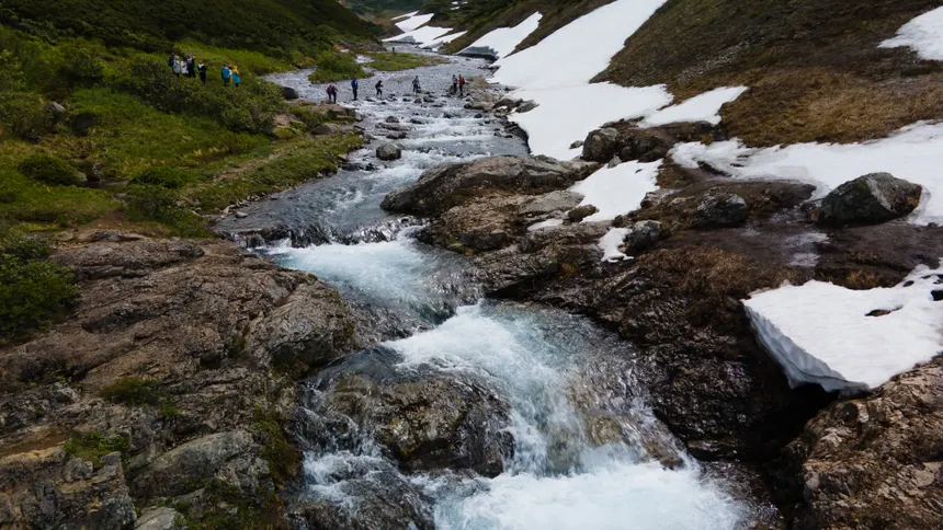A breathtaking view of a flowing mountain stream surrounded by snow patches and hikers in Kamchatka.