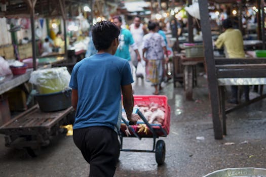 A man pushes a cart filled with goods through a bustling outdoor market, surrounded by shoppers and vendors.