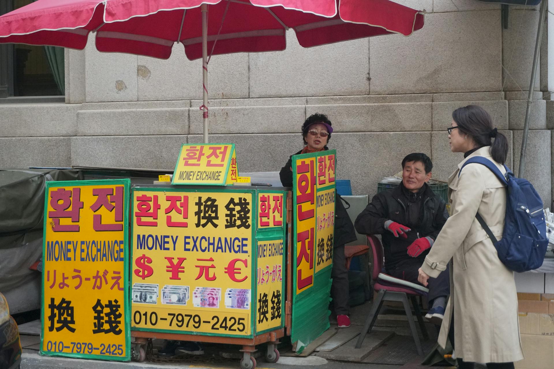 Two adults conversing at an outdoor money exchange booth with vivid signage in a city street.