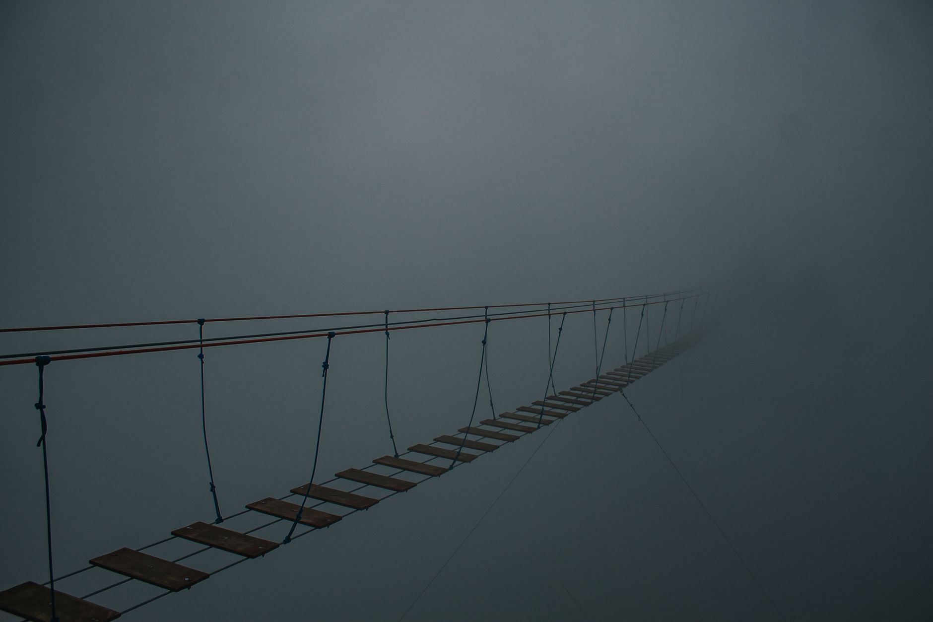 Spooky suspension bridge vanishing into a dense fog, creating a mysterious atmosphere.