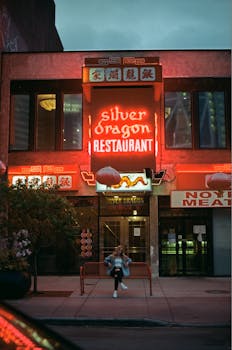 A person sits under neon lights at Silver Dragon Restaurant's entrance at night.