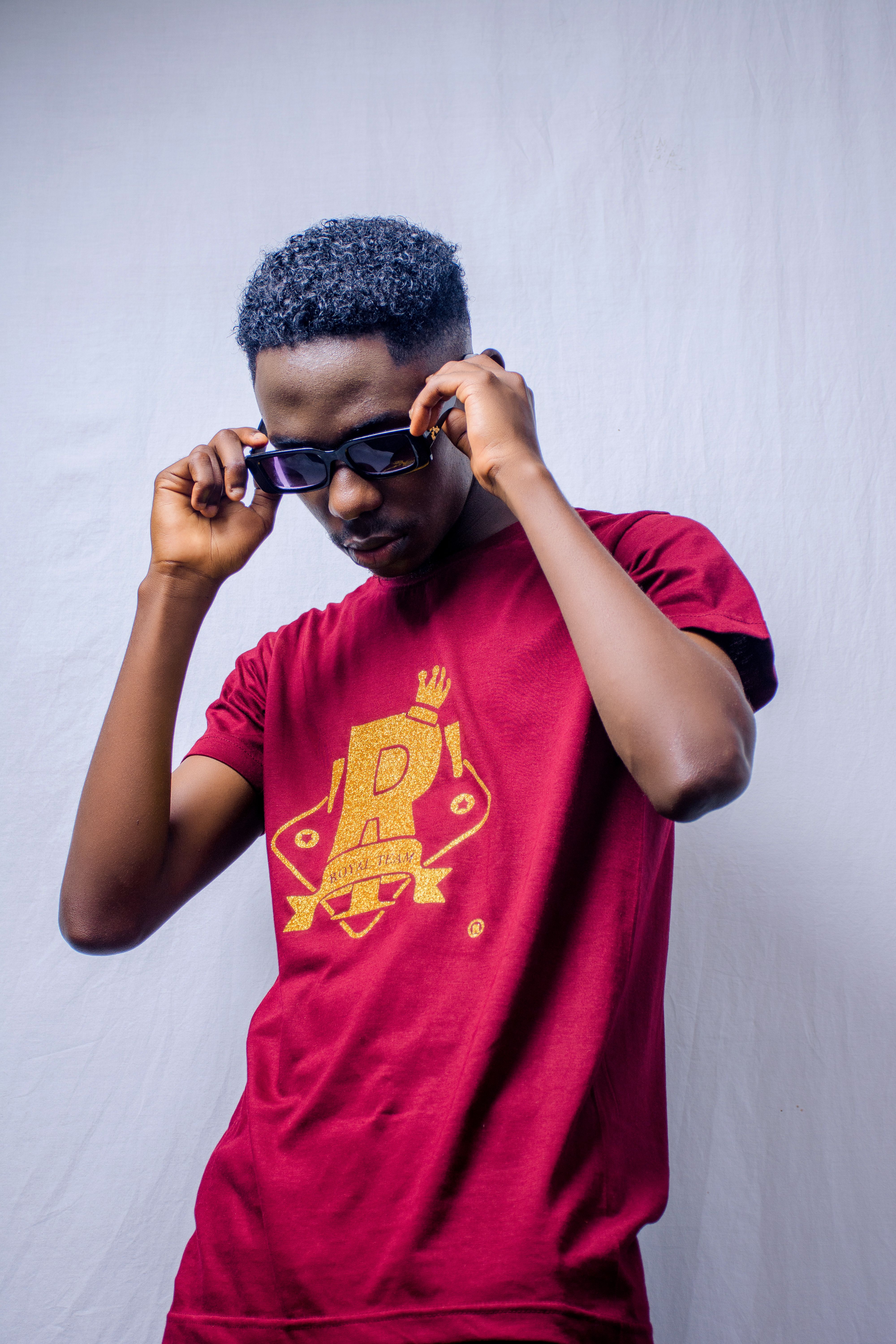 A fashionable young man adjusts his sunglasses, wearing a branded burgundy t-shirt in a studio setting.