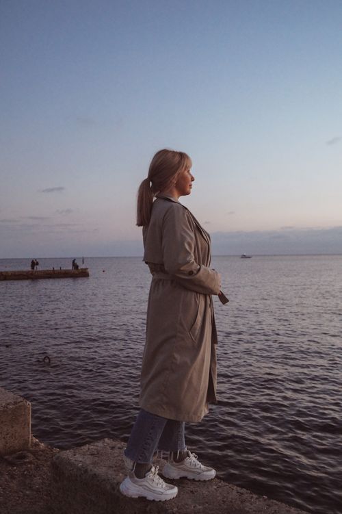 Free A woman in a trench coat stands on a concrete ledge, gazing at the sea during sunset. Stock Photo
